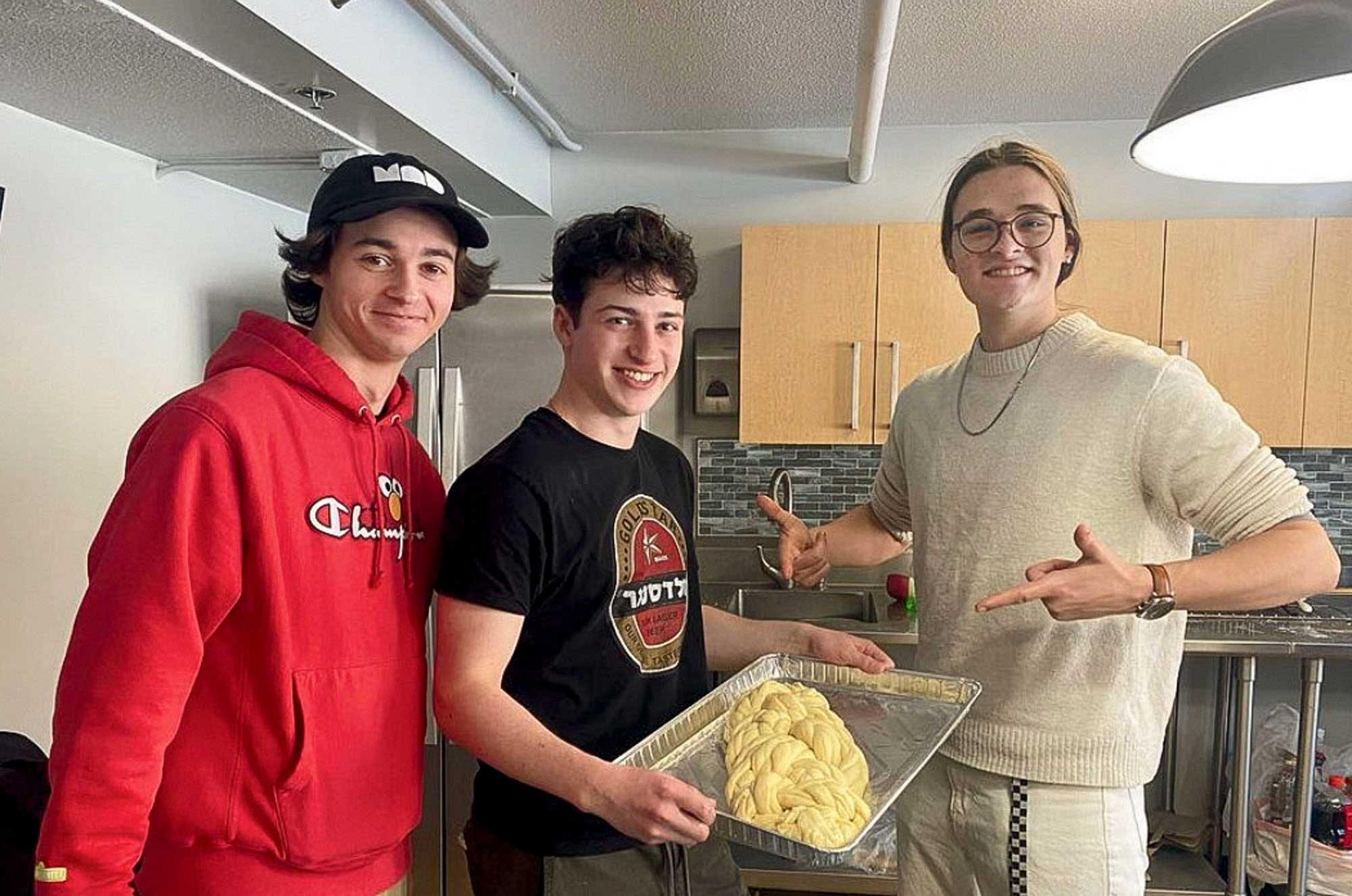 Three individuals are in a kitchen setting. The person on the left is wearing a red hoodie and a black cap, the middle person is wearing a black t-shirt with white hbrew text and is holding a baking tray with braided challah dough, and the person on the right is wearing a beige sweater and gesturing towards the dough with both hands. The background includes kitchen cabinets and appliances, suggesting a domestic cooking scene.