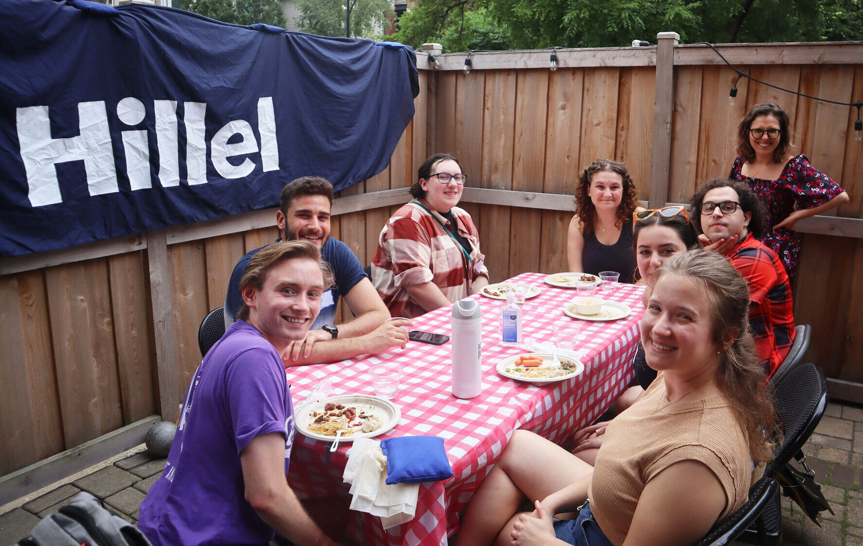 8 college students gathered around a table with red and white checkered table cloth eating dinner. The table is located in a backyard that is enclosed with brown wooden fence and blue flag that says "Hillel".