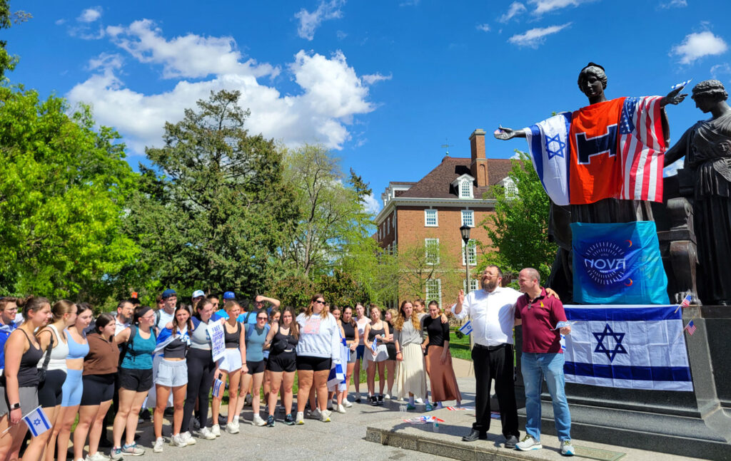 A group of college students with their arms around each other standing in front of a rabbi and another man as well as a statue draped with the American flag, two Israeli flags, a University of Illinois flag and a nova music festival flag.