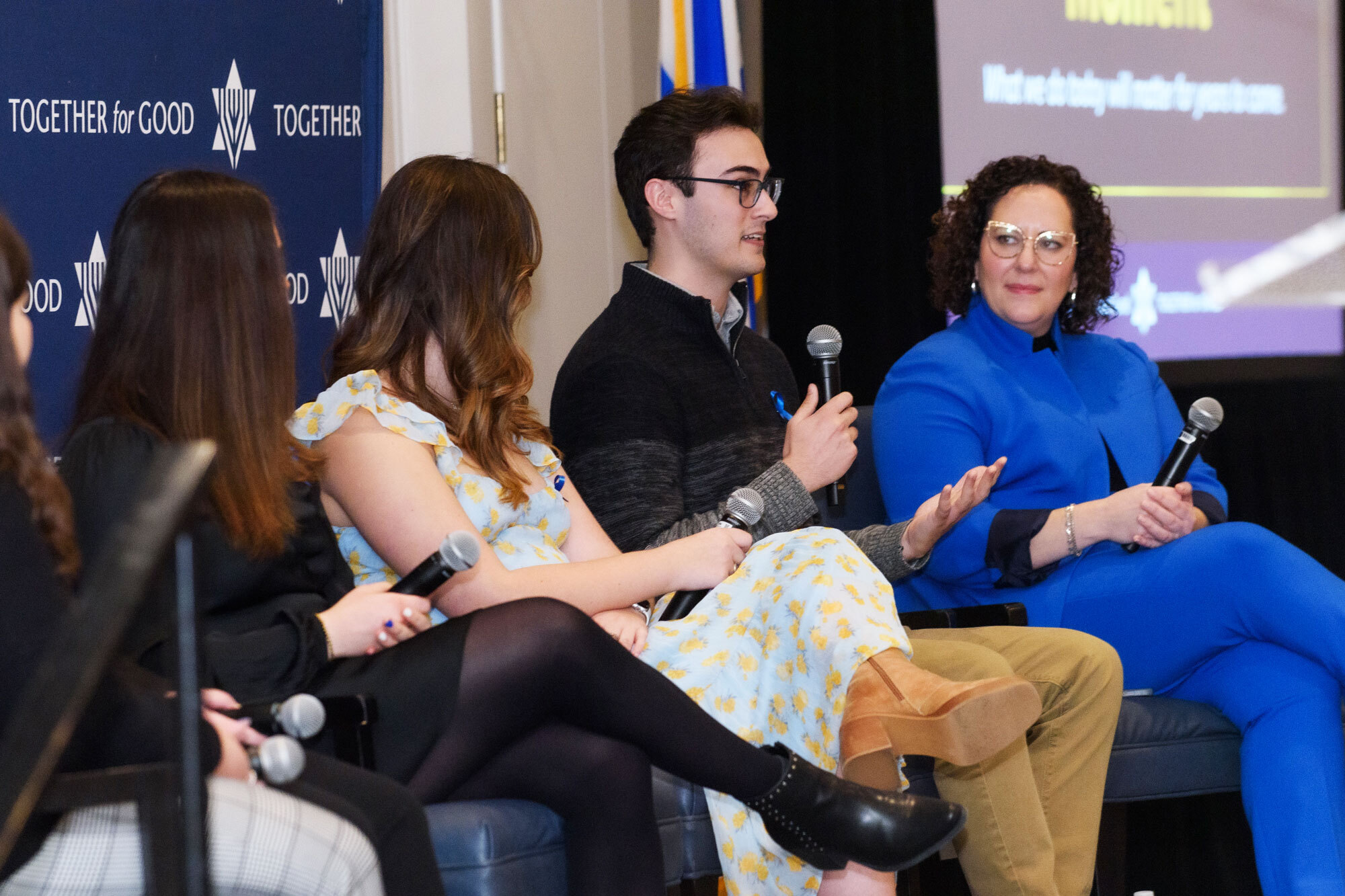 A JUF panel with four women and one man (who is talking) all wearing blue ribbon pins and holding microphones.