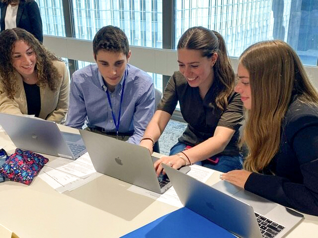 Four college students including three women and one man in business clothes gathered around three laptops.