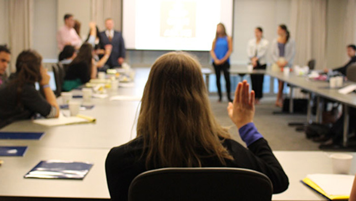 A woman in the forefront of the frame with her back turned to us and her hand raised to ask a question. This takes place in a conference room with a Rectangle shaped set of tables and a screen in the background.