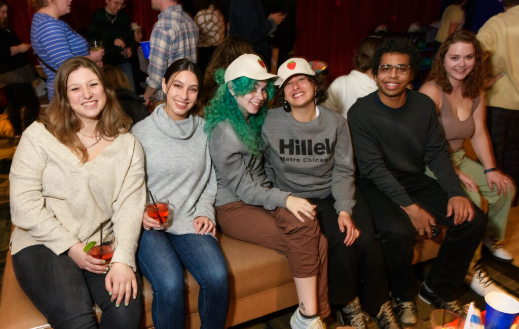 A group of six college students sitting on an orange bench indoors. The woman in the middle is wearing a 'Hillel' crewneck.