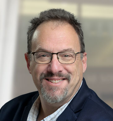 VP of COE Aaron Scott head shot of him smiling with glasses and a blue suit.