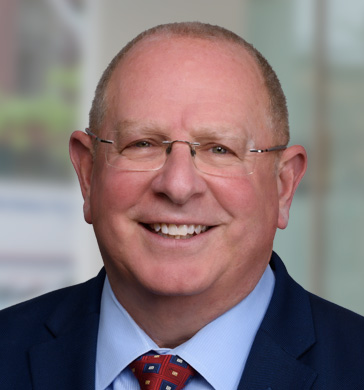 Executive Vice president Jay Tcath headshot smiling with glasses, a red tie and blue suit.