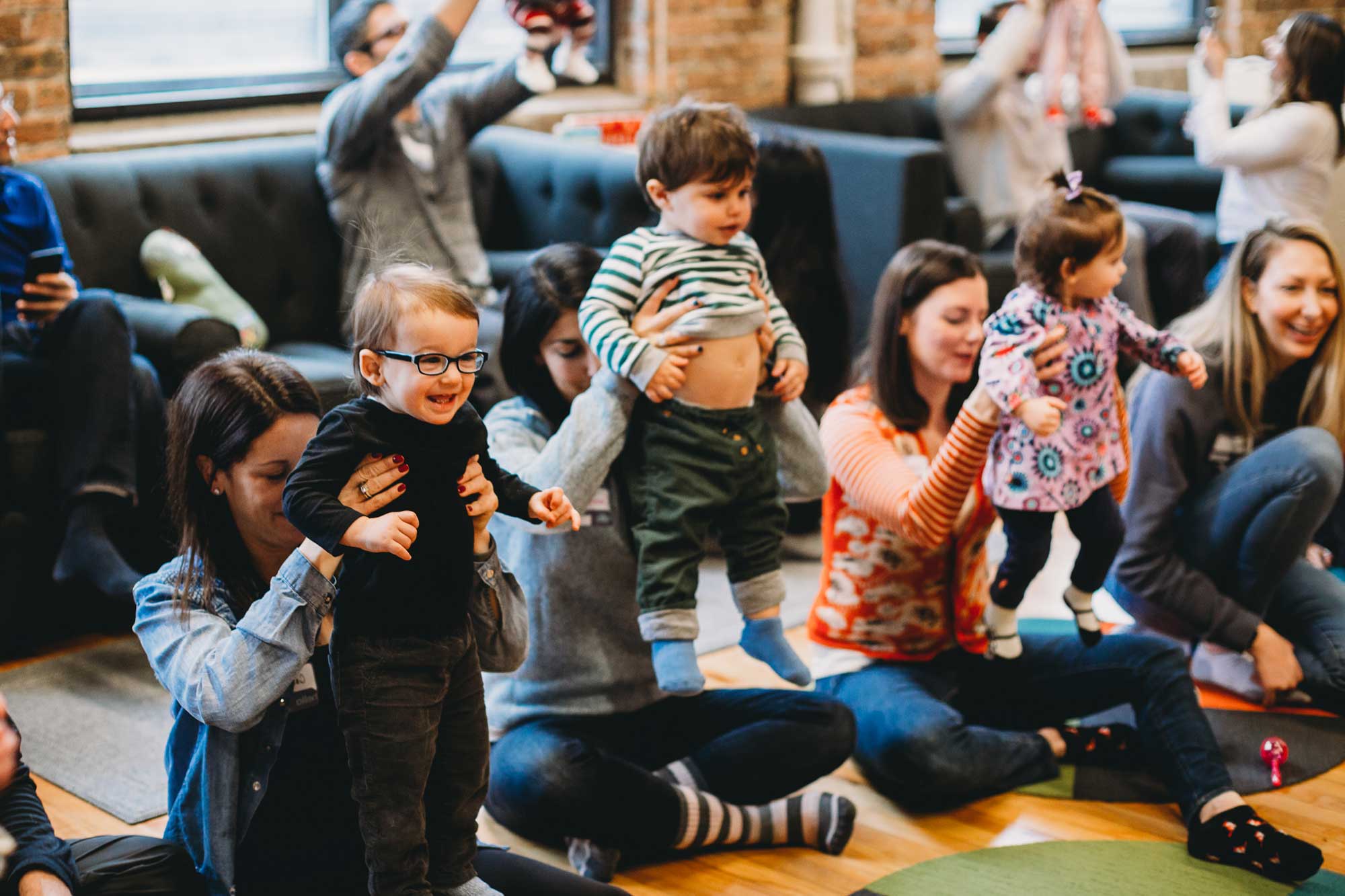 A group of individuals, including several children and adults, are gathered in an indoor playroom. The room features wooden floors and large windows that allow natural light to flood in. Some children are standing, while others are being held by adults. The adults appear to be engaged in conversation or play with the children. The setting is casual and welcoming, with various toys and play equipment visible in the background.