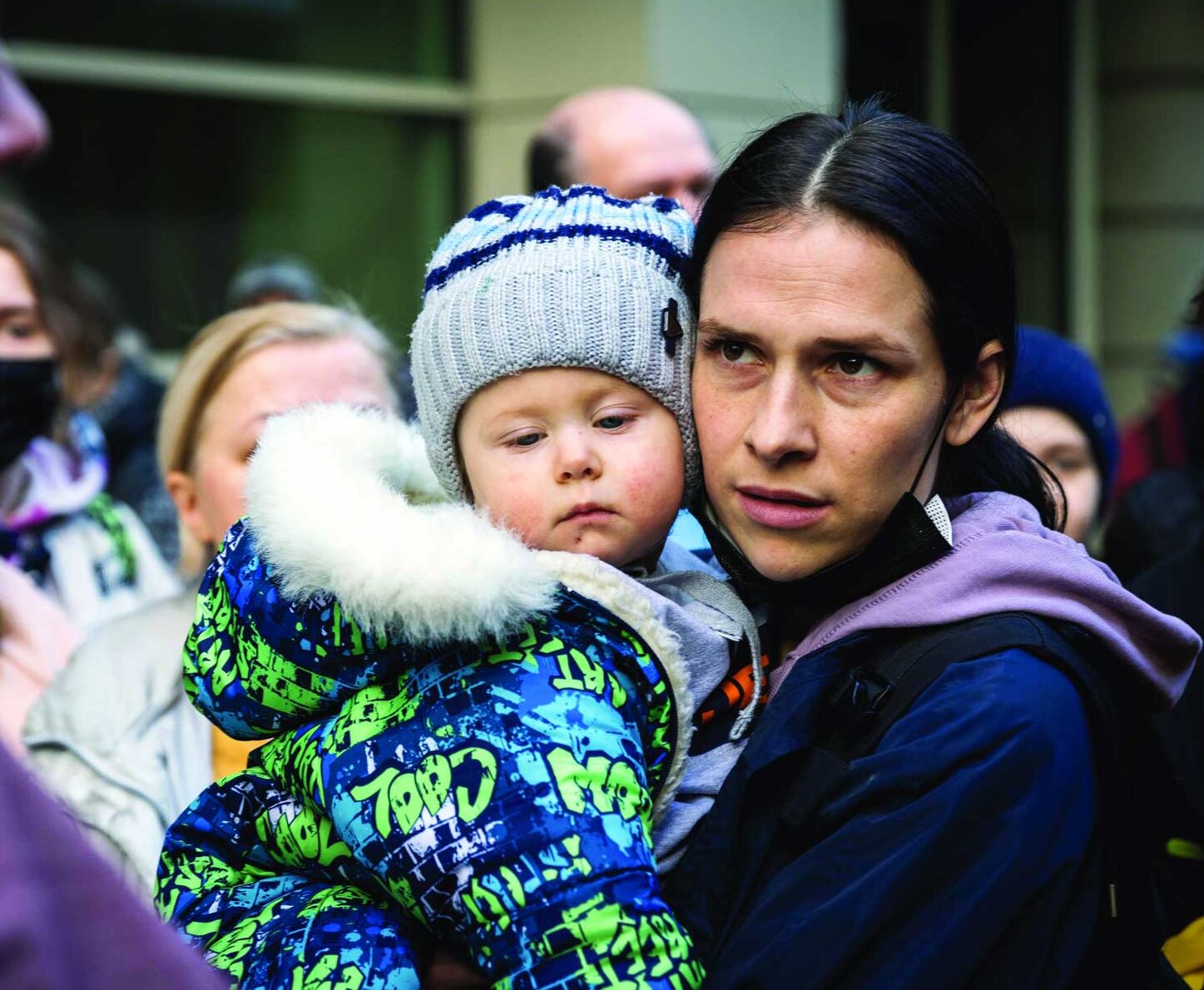 A woman holding a small child wearing a blue and green pattered winter jacket and a grey beanie.
