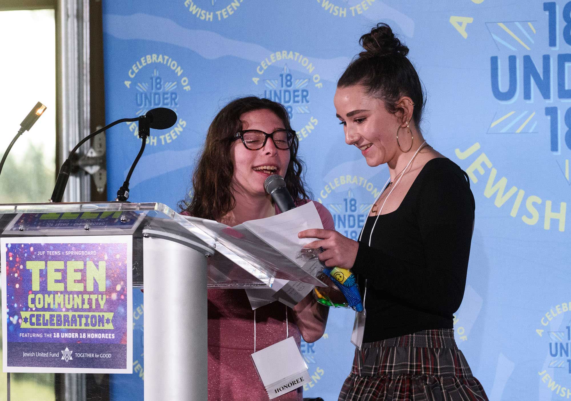 Two individuals with obscured faces stand at a podium with a microphone, during what appears to be a public speaking event. One individual is holding papers, possibly preparing to speak. The backdrop features the words “TEEN COMMUNITY CELEBRATION” along with various decorative elements such as stars and lines. The podium has a sign that reads “18 UNDER 18 JEWISH”.