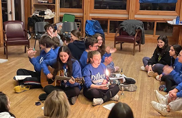 A group of teens sitting in a circle on the floor with a guitar and Havdalah candle singing.