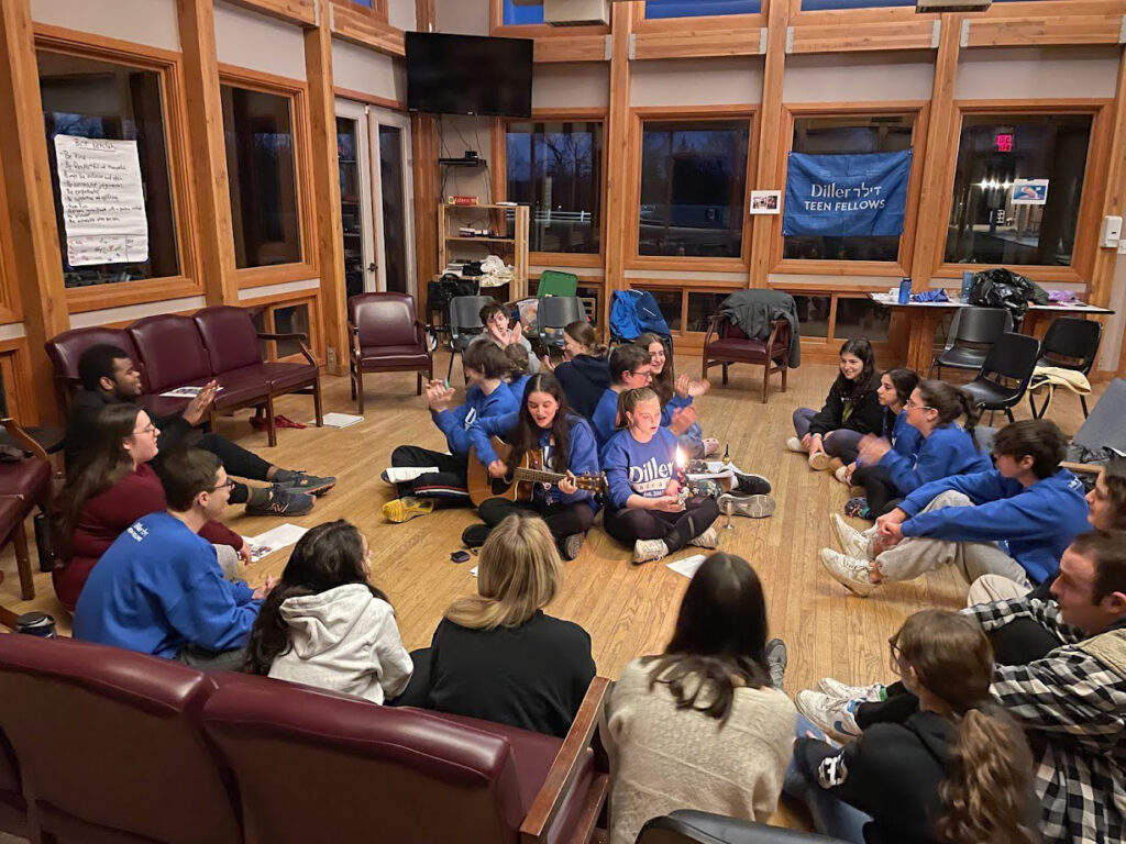 A group of teens sitting in a circle on the floor with a guitar and Havdalah candle singing.