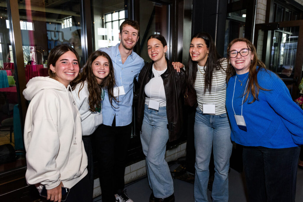 A group of six teens smiling with arms around each other each wearing white name tags necklaces.