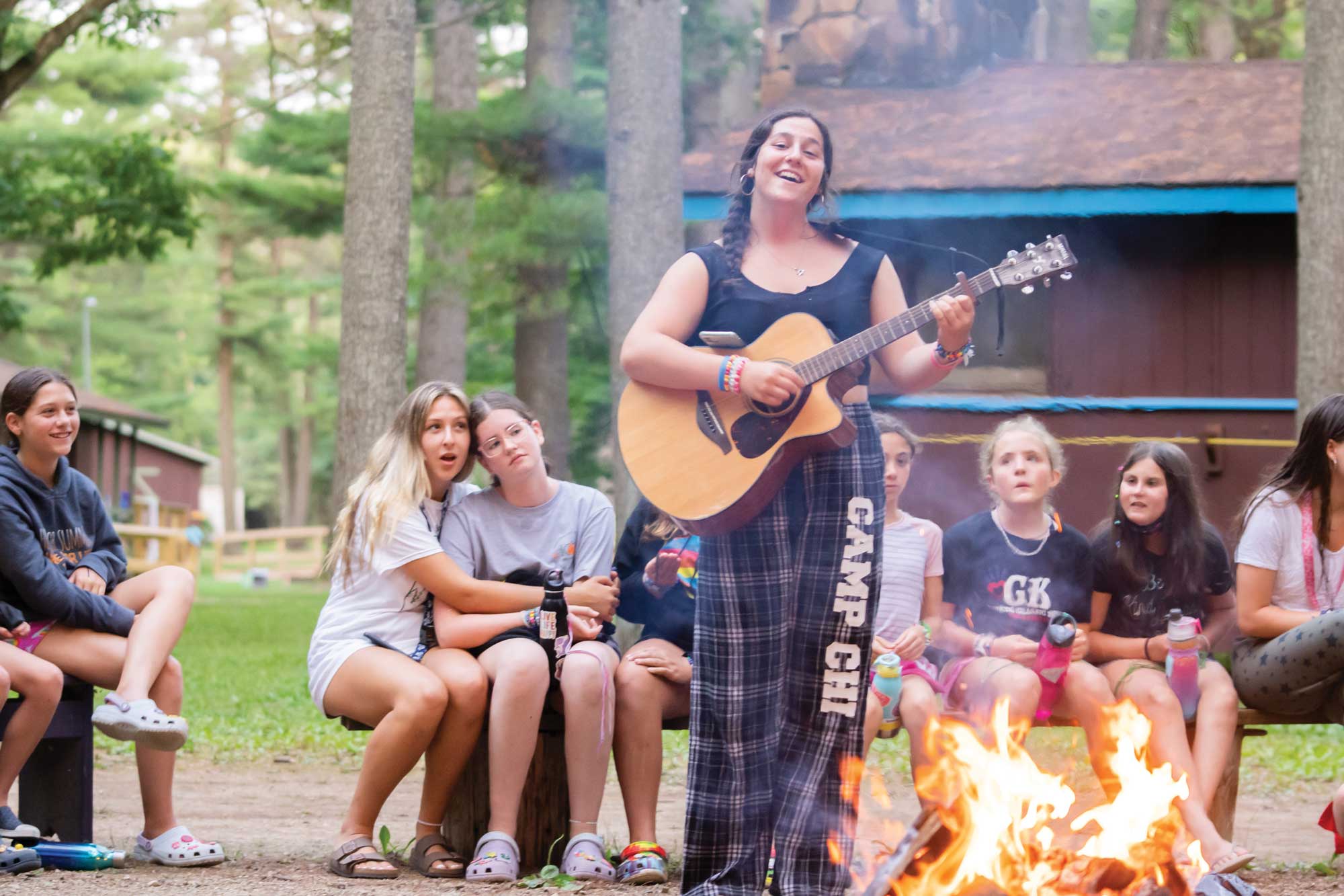 A teen girl wearing plaid pants reading "Camp Chi" singing with a guitar for a group of campers around a fire pit.