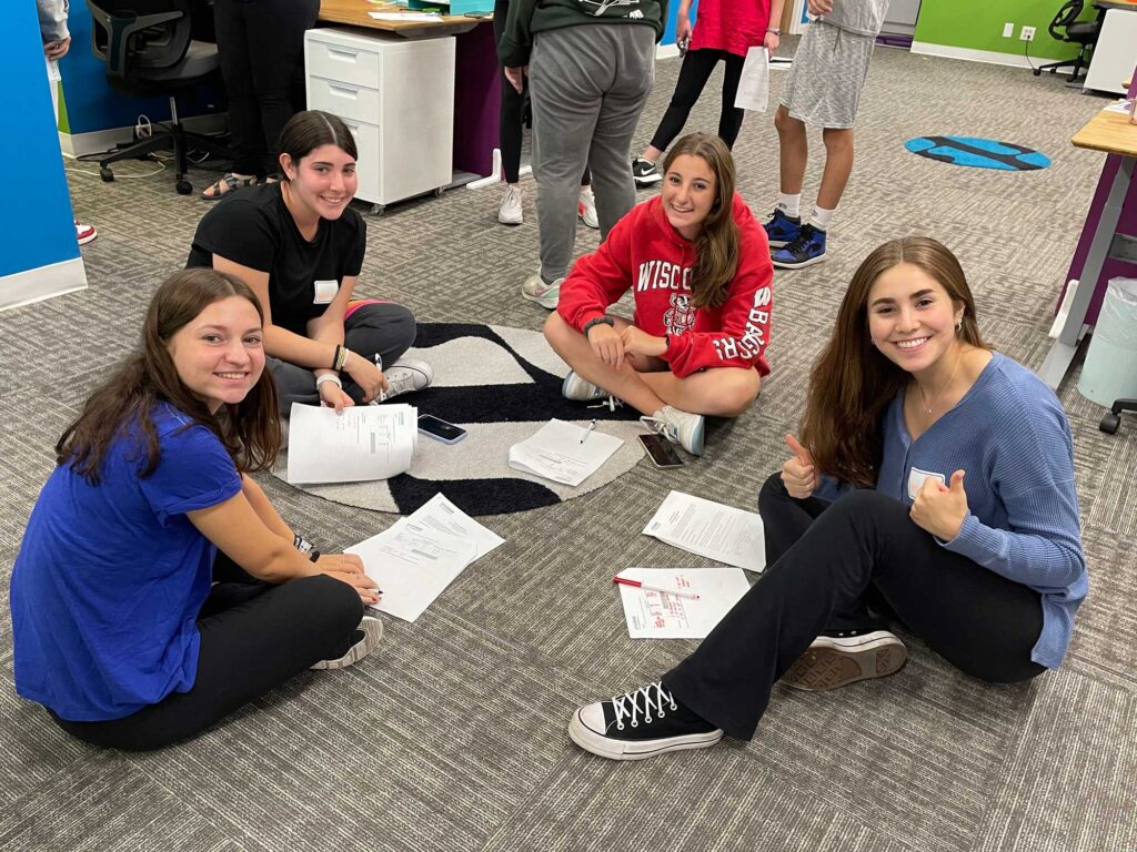 Four springboard ambassadors sitting on a carpeted floor smiling with thumbs up while wearing name tags and using pen and paper to fill out their discussion page.