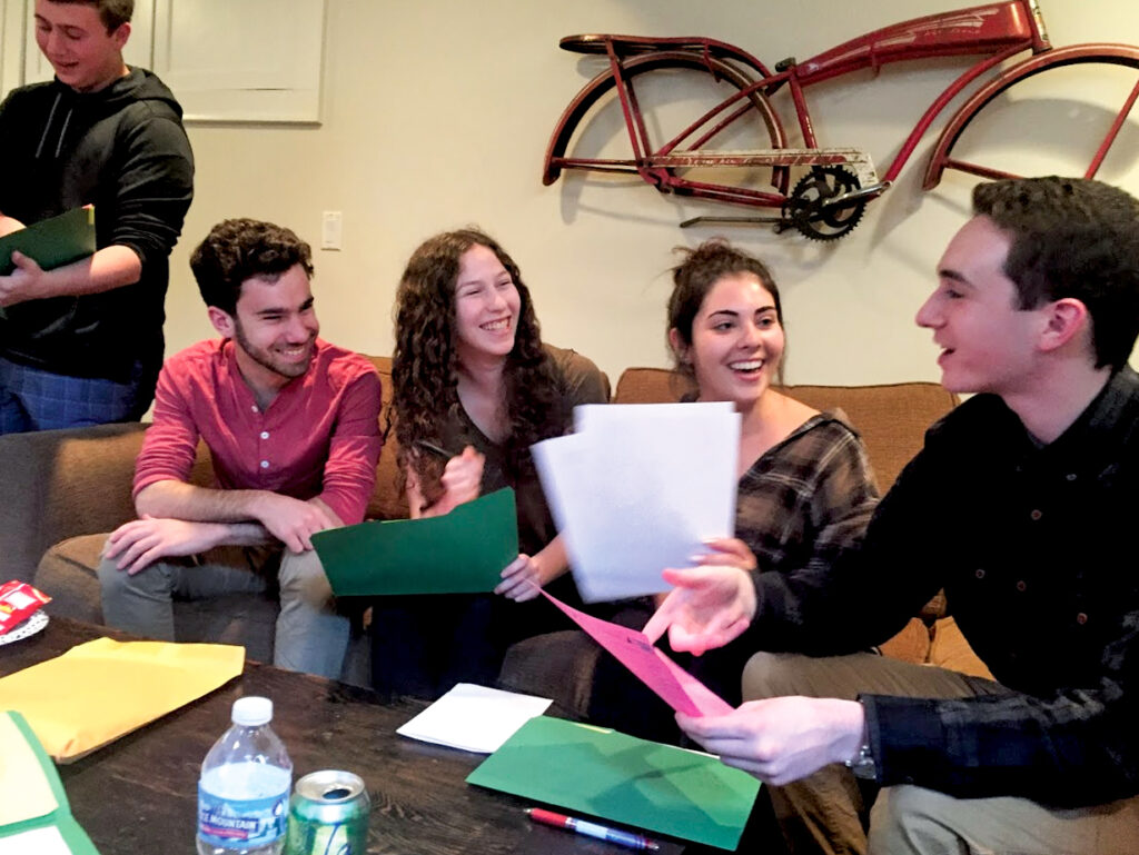 A group of four teens sitting on a couch with green folders laughing and discussing a paper.