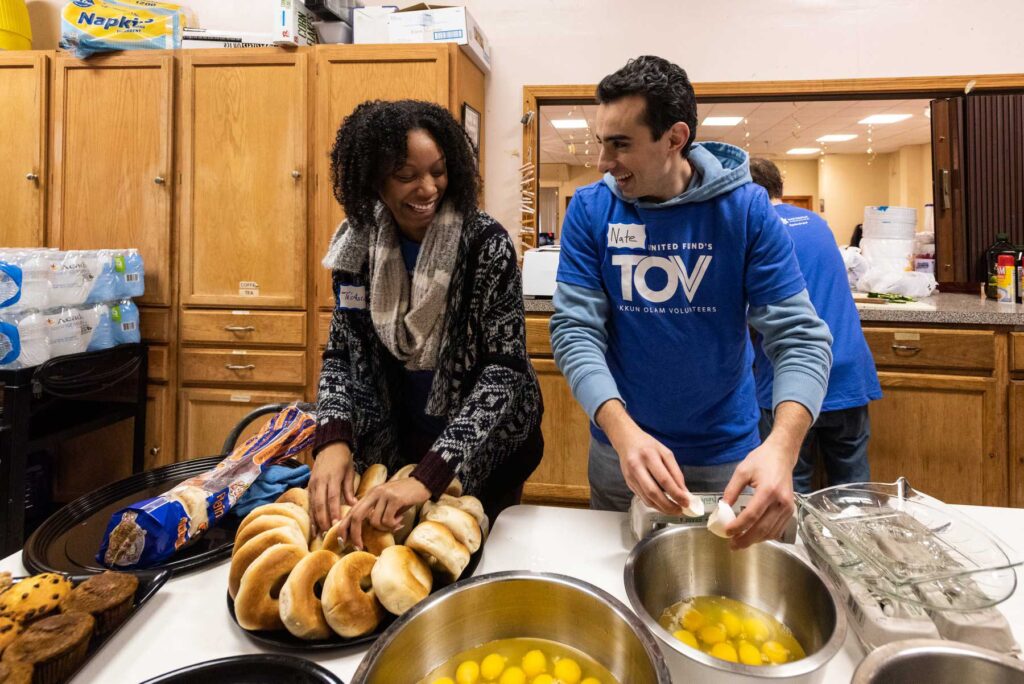 A man and a woman in a kitchen volunteering with bagels while cracking eggs and wearing TOV volunteering shirts.