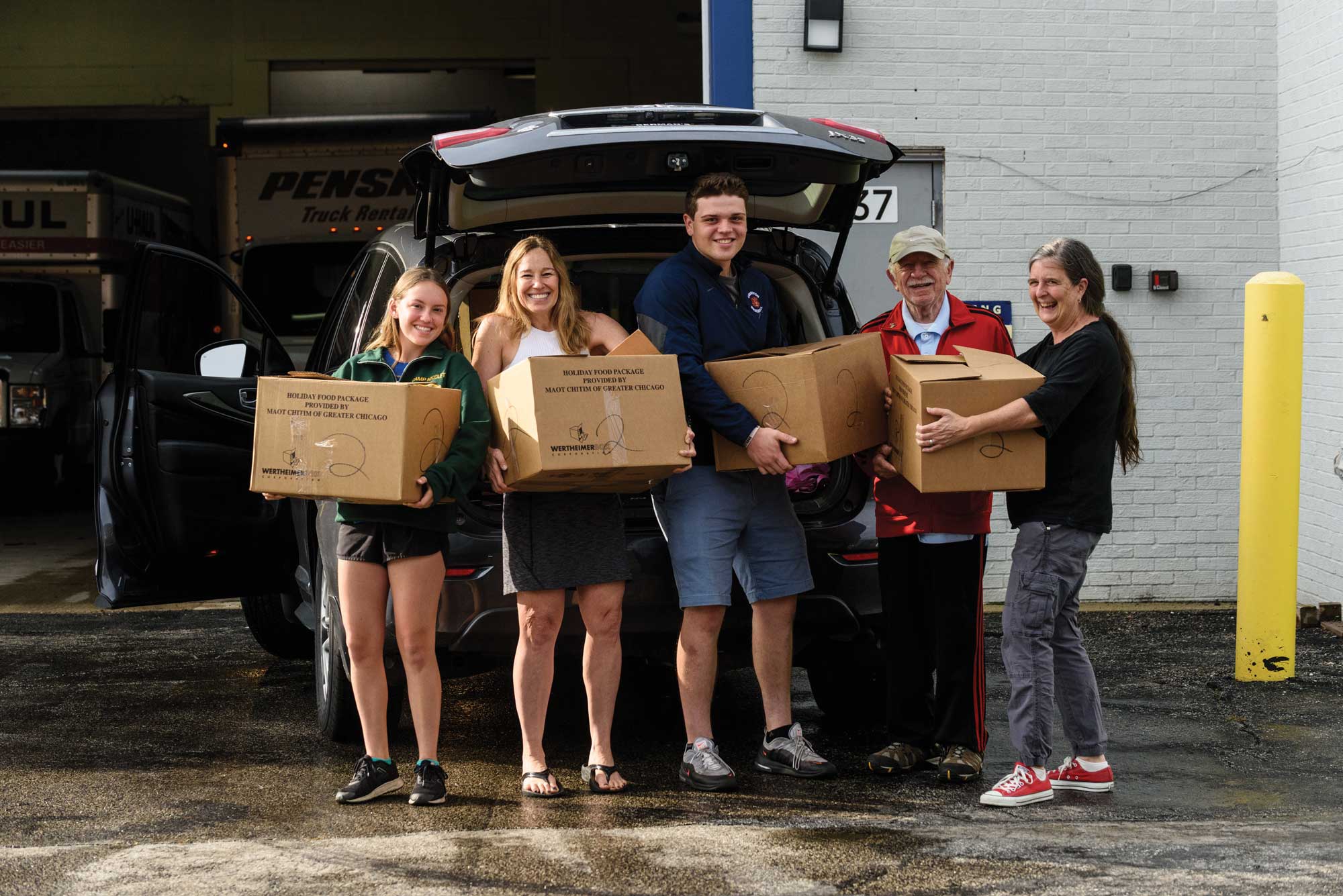 Five adults volunteering while holding brown cardboard boxes outside of a car with a trunk open.