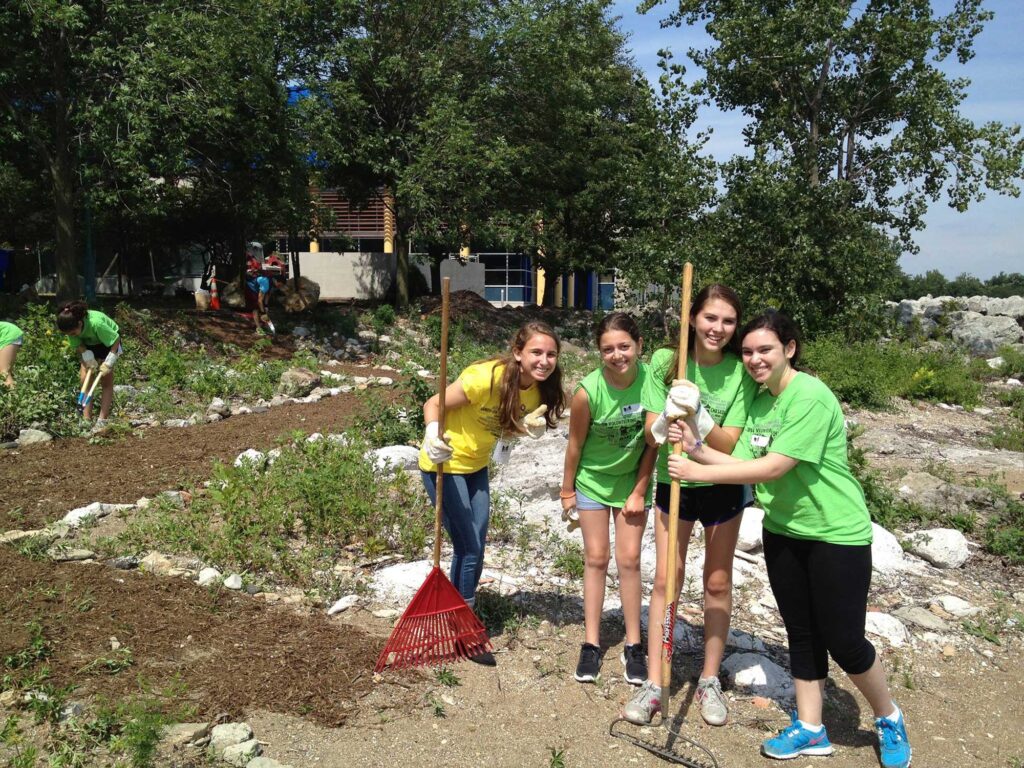 Four teen girls standing in a garden volunteering with rakes.