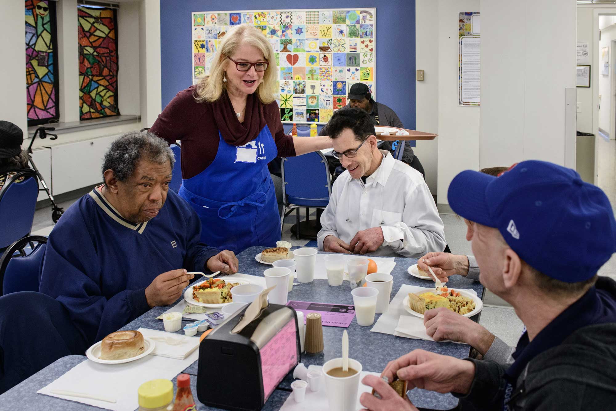 A group of adults sitting at a table eating with a JUF Uptown cafe volunteer serving the guests.