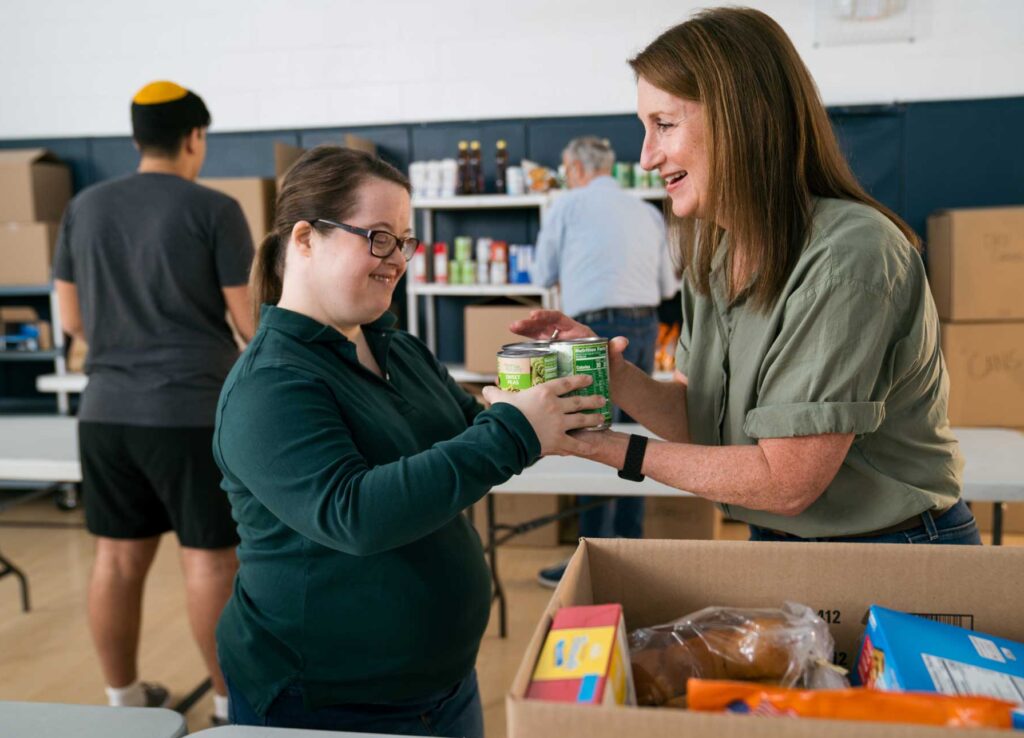 One individual is handing a can to the other, and there are boxes on a table that suggest some sort of food distribution or collection event.