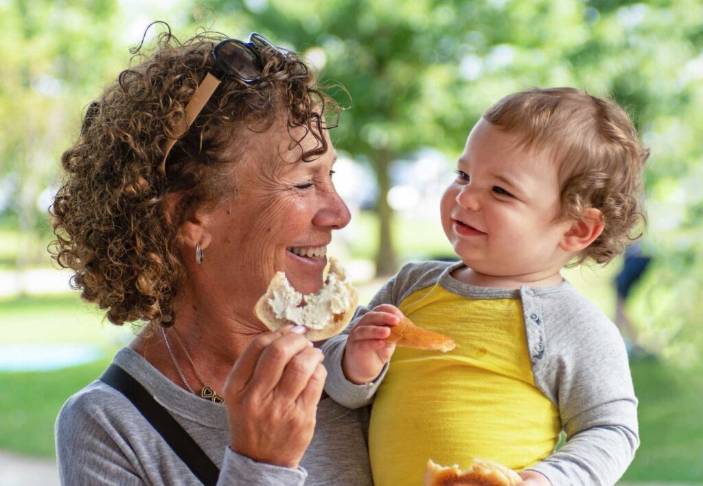 An adult holding a bagel with cream cheese in one hand and a baby in the other arm.