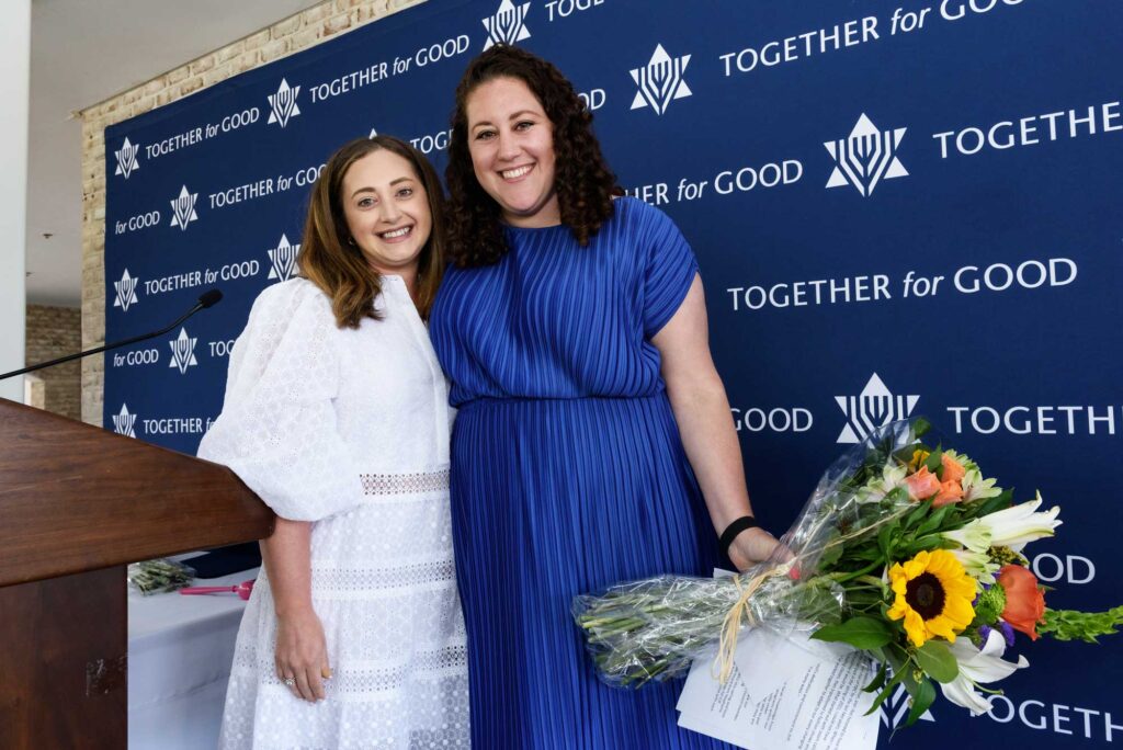 Two women smiling in front of a JUF "Together for Good" branded back drop where one woman is wearing a blue dress holding a bouquet of flowers and the other wearing a white dress.