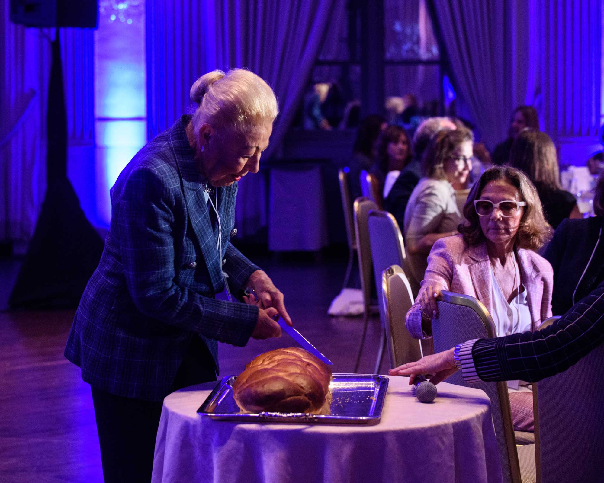 An older woman cutting as loaf of challah at a formal event.