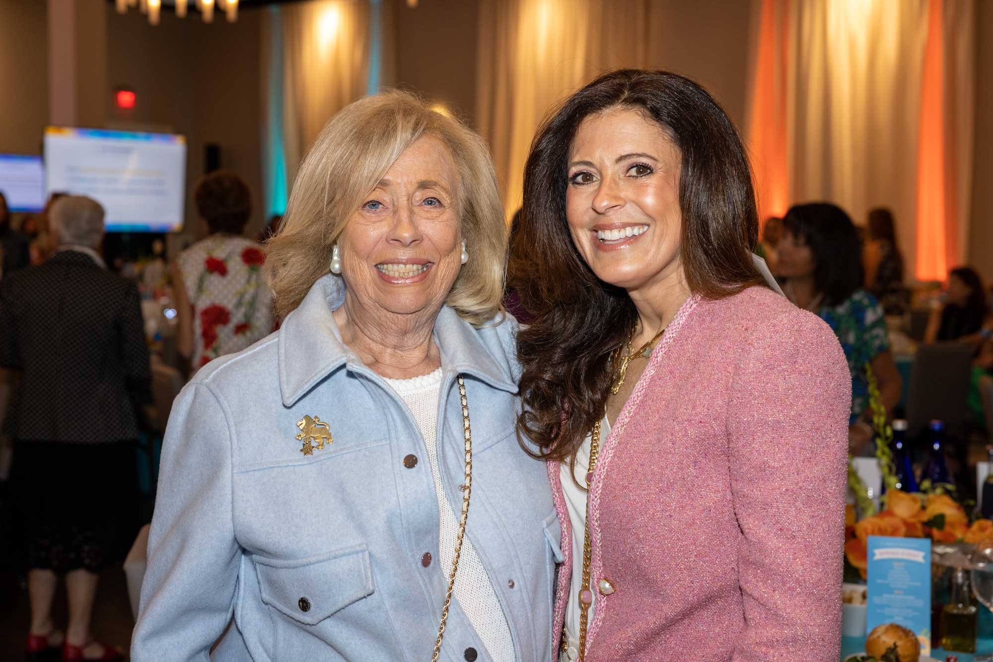 Two women smiling for the camera wearing blue and pink blazers and "lion of Judah" pins at a women's giving event.