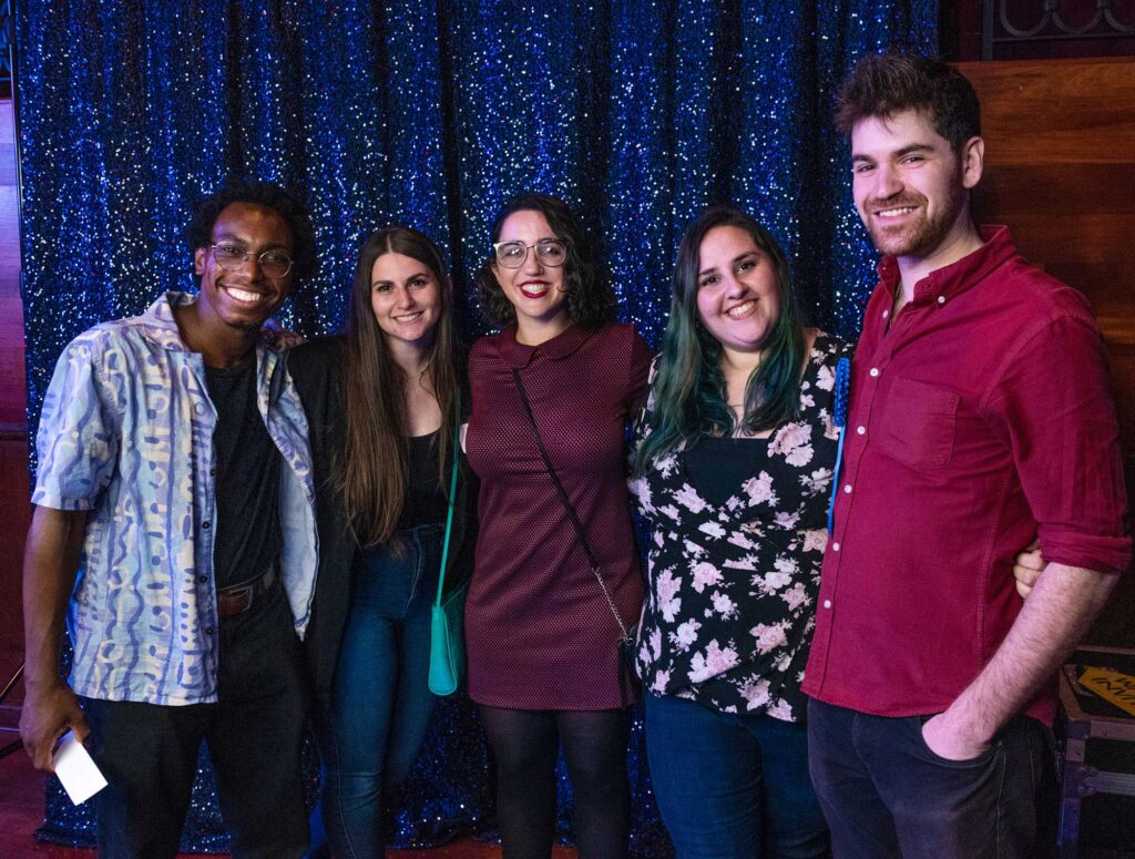 5 young adults standing in front of a dark blue sparkly curtain. From left to right: a black man with glasses wearing a blue open button up collared shirt, a brunette woman with long hair wearing a teal purse, a black haired woman with glasses and a maroon shirt, a  woman wearing a black and white floral shirt with teal hair and a tall brunette man wearing a red button up collared shirt with a pocket.