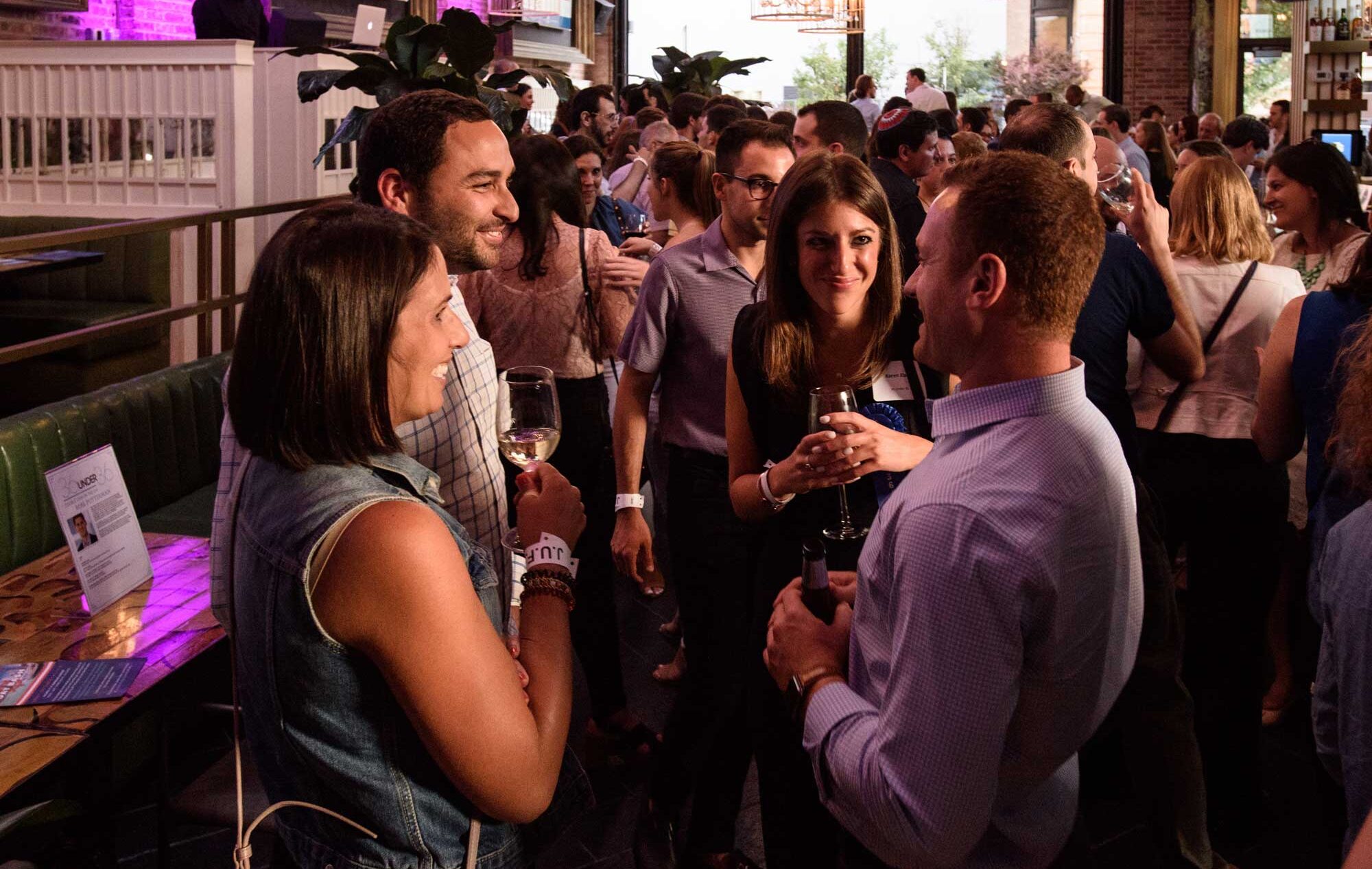 Four young adults smiling holding wine glasses and beer bottles in a bar networking event including two men and two women.