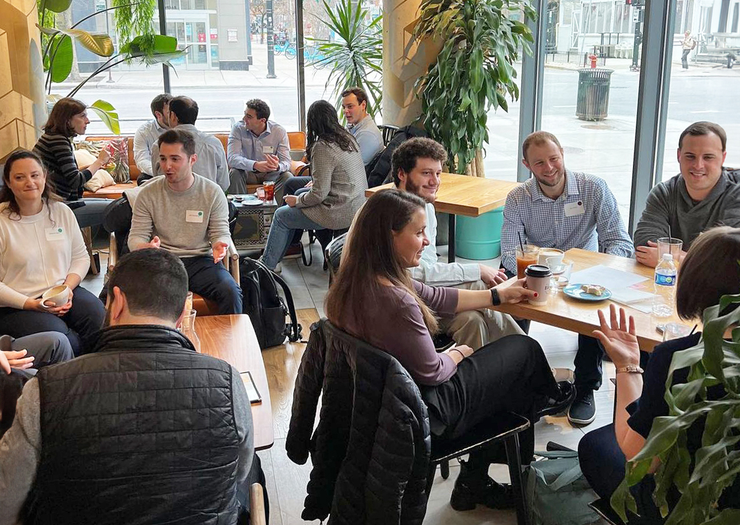 Young adults partaking in professional development in small groups sitting at various tables at a coffee shope wearing name tags and having discussion.