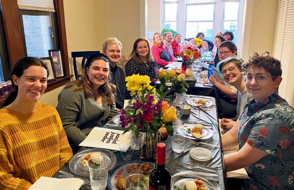 A group of young adults sitting and smiling at a long seder table with floral arrangements and seder plates displayed.