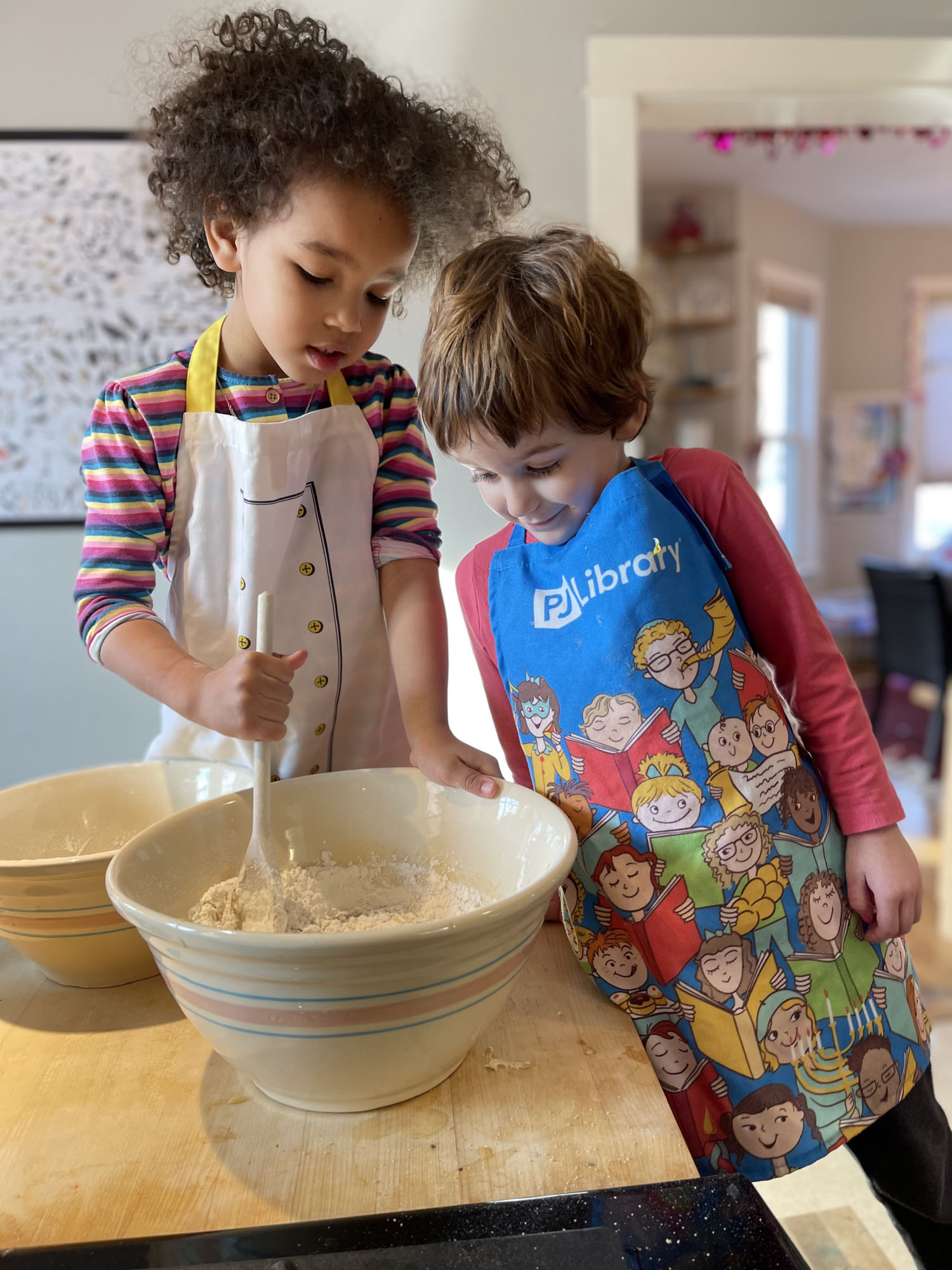 Two young toddlers mixing flower in a mixing bowl together while one wears an apron that says "PJ Library" on it.