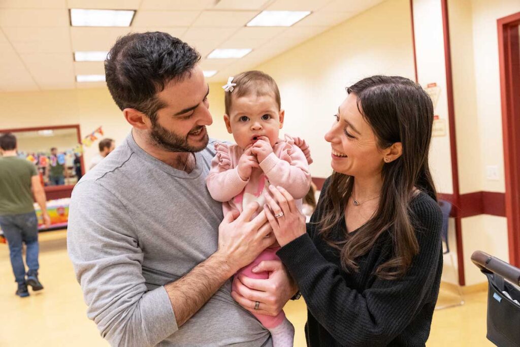 A mom and dad holding their baby girl in a pink outfit with a bow while staring at her in the middle of them.