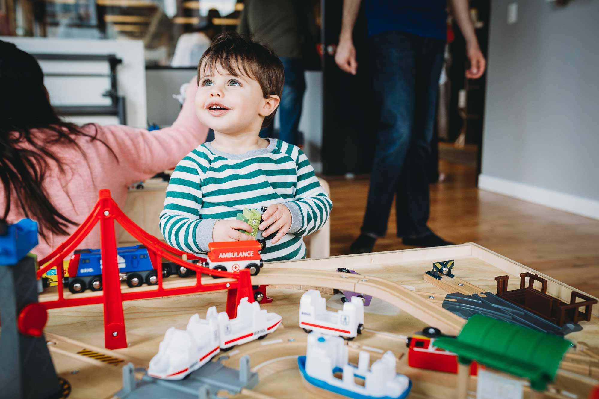 A toddler boy holding a train while looking up and standing in front of a train table.