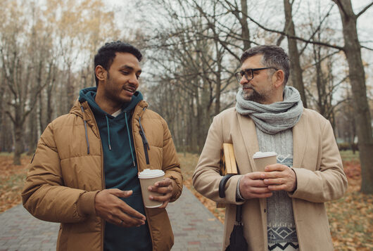 Friends, a senior and a young man walking and talking and drinking coffee together in the autumn park.
