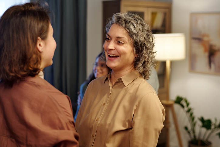 Cheerful mature woman in beige blouse laughing and looking at her daughter while greeting her in the corridor of apartment.