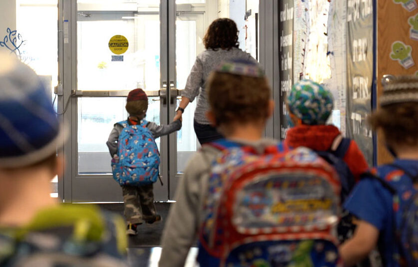 A preschool teacher holding hands with a toddler wearing a kippah as many toddlers in kippah's exit the preschool building.