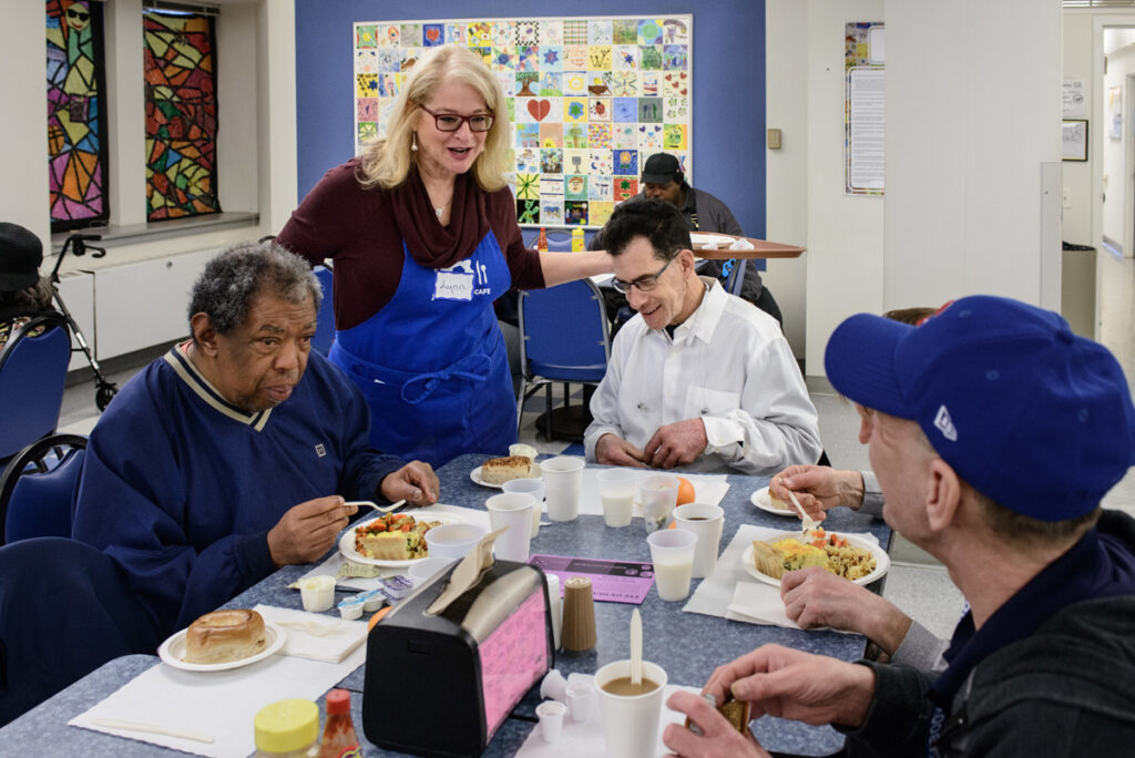 Three older men sitting at a communal table eating as an Ezra volunteer greets them.