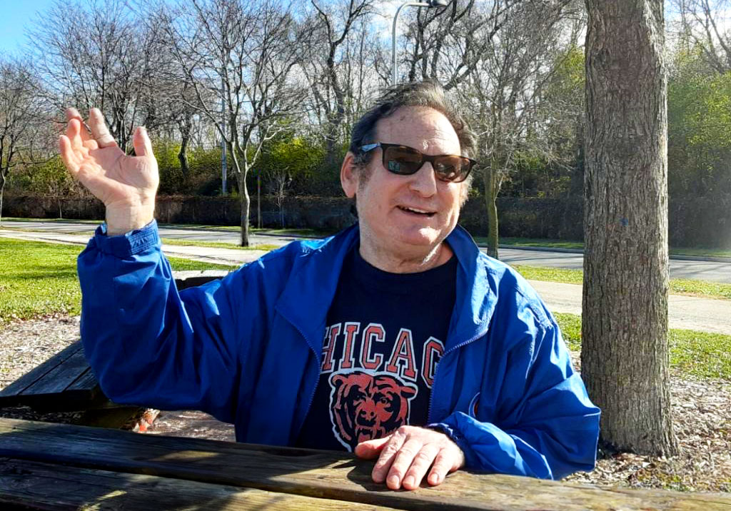 An older man with a disability sitting at a picnic bench wearing a Chicago bears shirt and sunglasses.