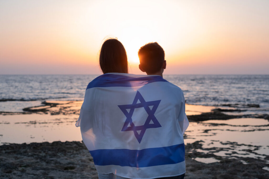 Two kids wrapped in an Israeli flag overlooking a sunset on a beach.