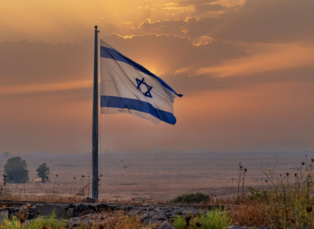 An Israeli flag flying in the wind with a sunset in the background.