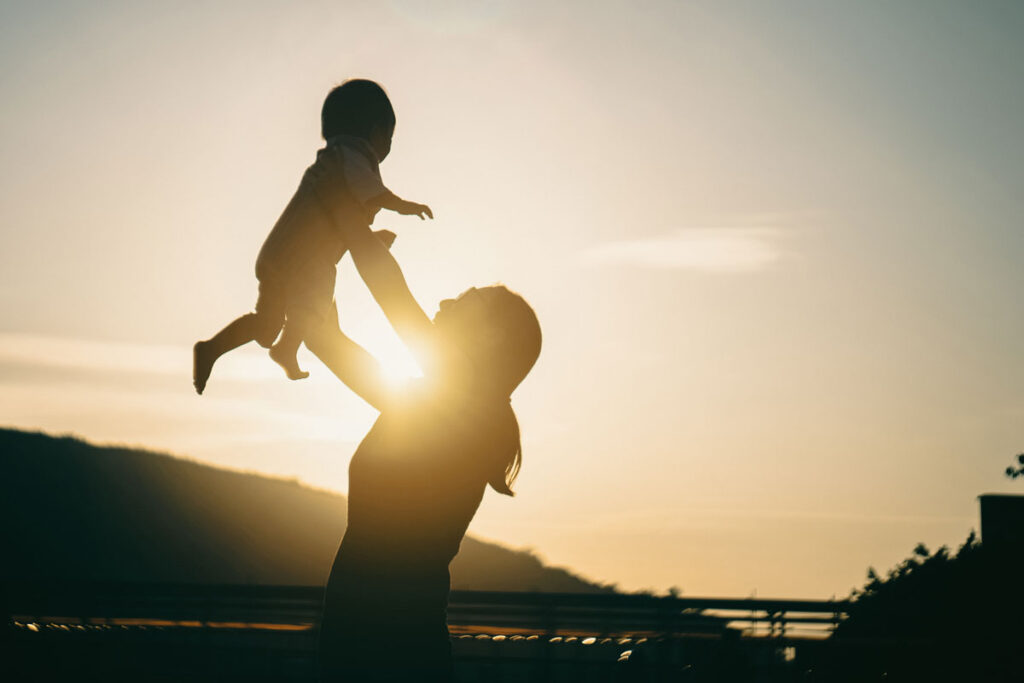 Silhouette of a mother holding up in the air her baby with beams of sunlight coming through.