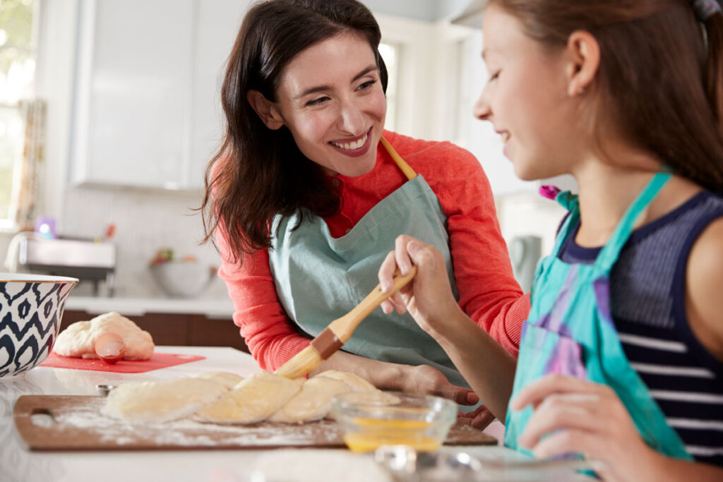 Woman in red sweater and blue apron smiling at young girl while making challah together