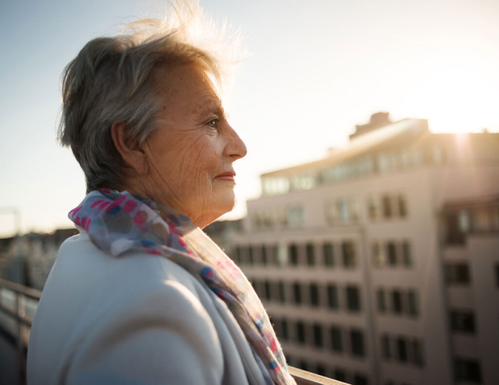 An older woman's side portrait looking off to the distance with hope on top of a building as sun beams fill the sky.