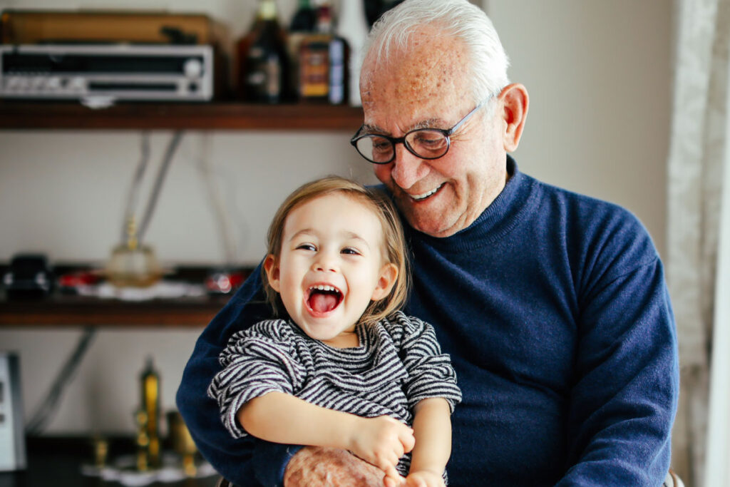 An old man with white hair wearing black glasses and a blue sweater holding his young granddaughter in his lap.
