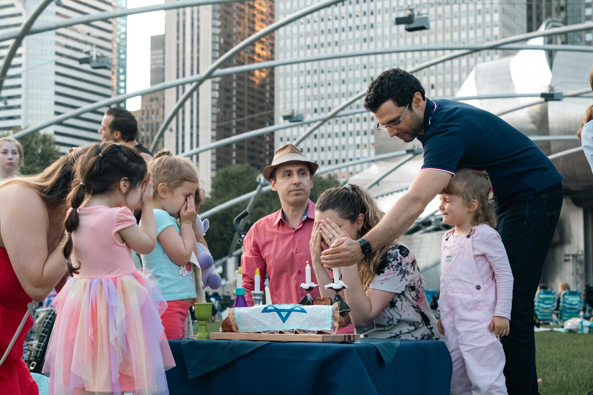 Three toddlers and one woman covering their eyes along side two men while blessing shabbat candles and a challah with a Jewish star over it outdoors.