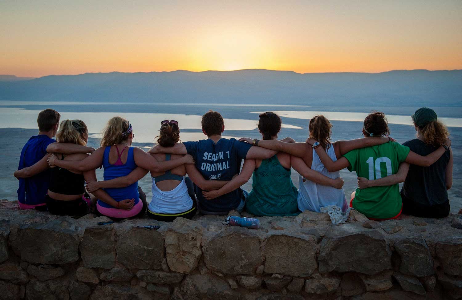 A group of young adults sitting on rocks with arms around each other facing the sunset in Israel.