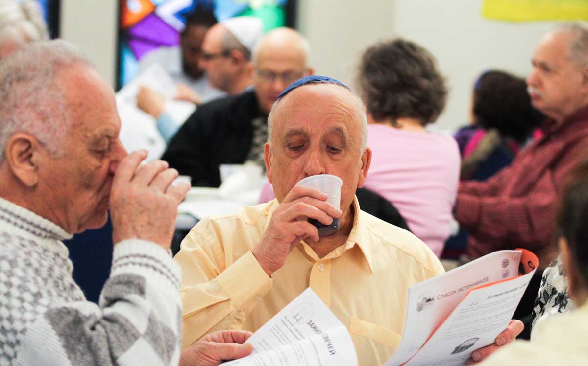 Two older Jewish men drinking red wine in plastic cups while reading prayers.