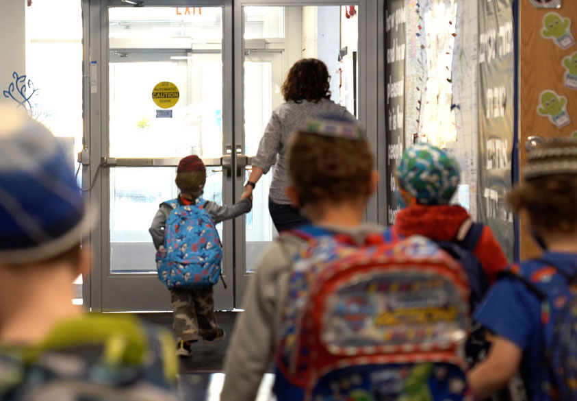 A preschool teacher holding hands with a toddler wearing a kippah walking out of the school followed by other young boys.