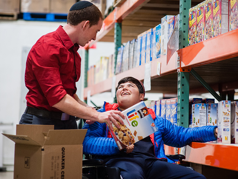 A man in a red shirt and a kippah handing a boy a box of cereal in a wheelchair while volunteering in a food pantry.